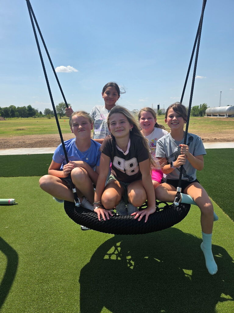 Five girls sit on a tire swing outside on a green field under a clear blue sky.