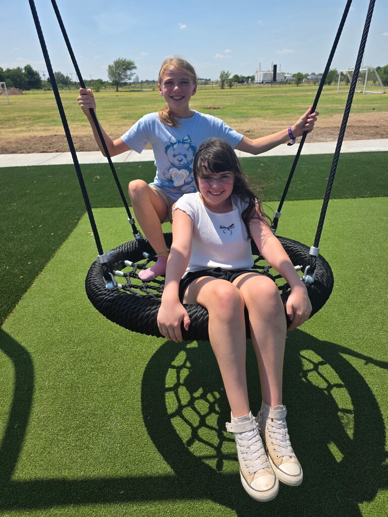 Two young girls sit on a tire swing at a park. One holds the ropes, and the other smiles.