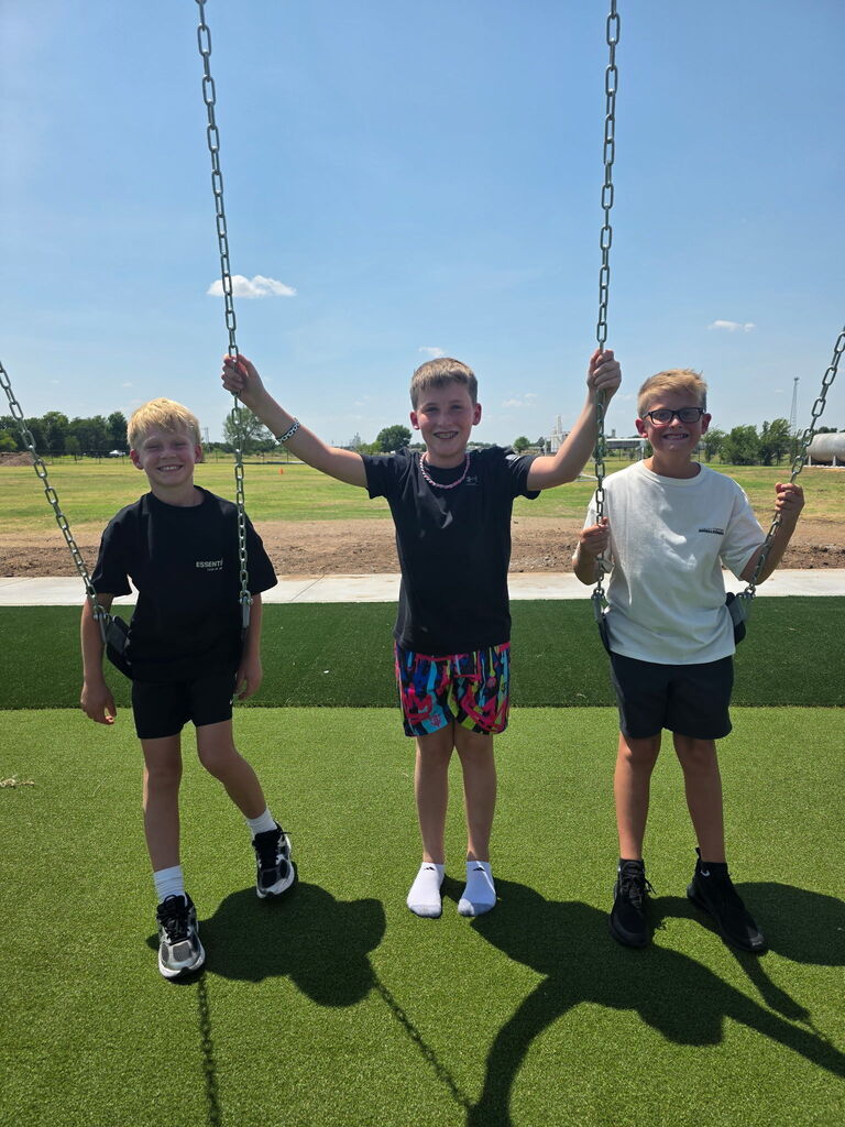 Three boys standing in an outdoor park, one standing on a swing with arms raised.