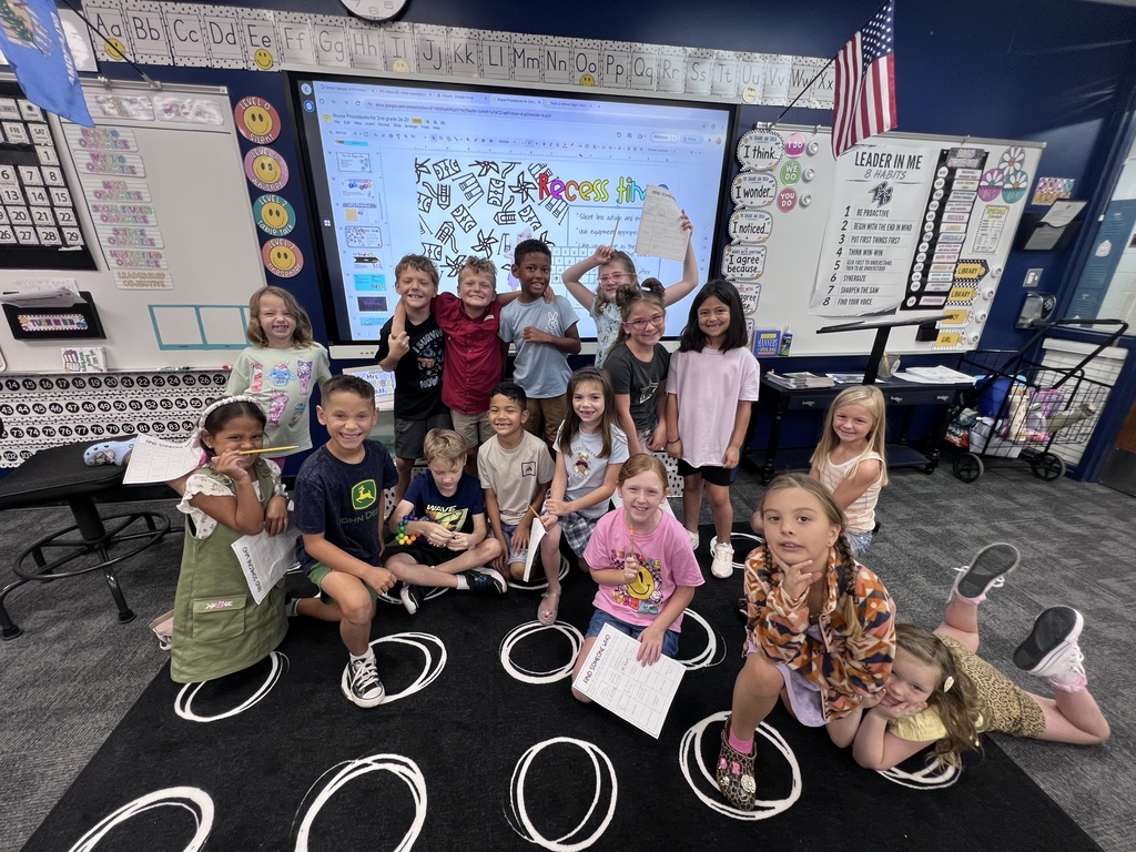 Group of young children in a classroom, standing and sitting, some holding papers, with a large screen and circle game in the foreground.