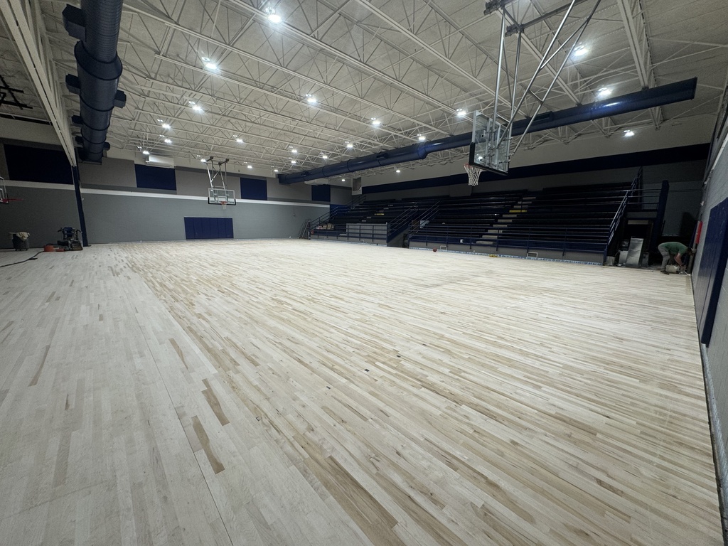 A large indoor court with polished hardwood floors. Overhead lighting and empty bleachers on one side.