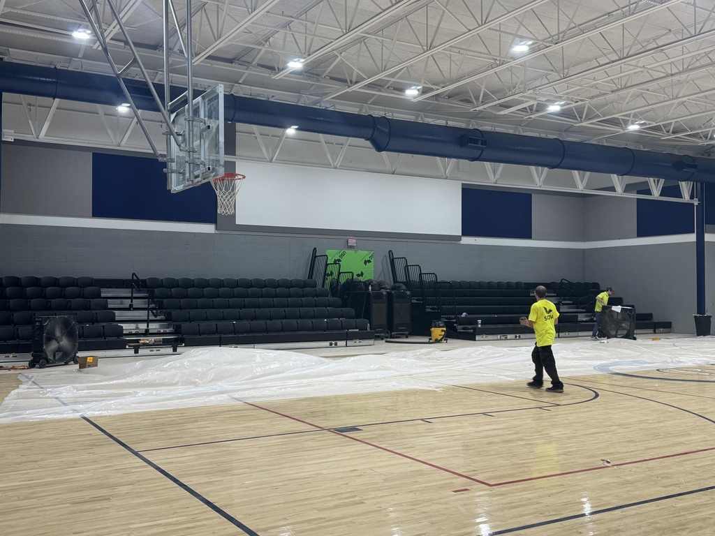 Indoor basketball court with wooden floor, empty bleachers, and workers cleaning. Two workers in yellow shirts are at the center.