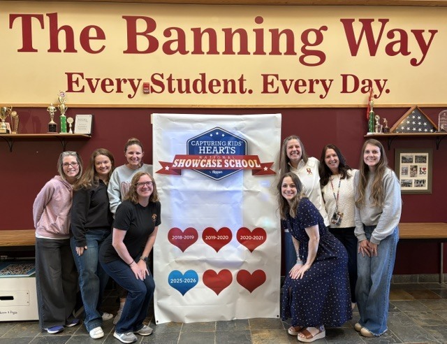 A group of eight smiling staff members pose indoors beneath a large sign reading, “The Banning Way – Every Student. Every Day.” They stand around a display celebrating the school's recognition as a CKH National Showcase School, featuring a banner with heart-shaped milestones and years of achievement. Trophy cases and school memorabilia are visible in the background, highlighting a welcoming school environment and team accomplishment.