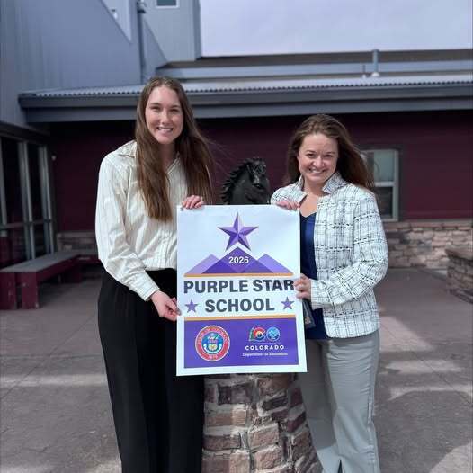 2 people standing outside holding a purple and white banner that says" purple star school" with government and Colorado emblems