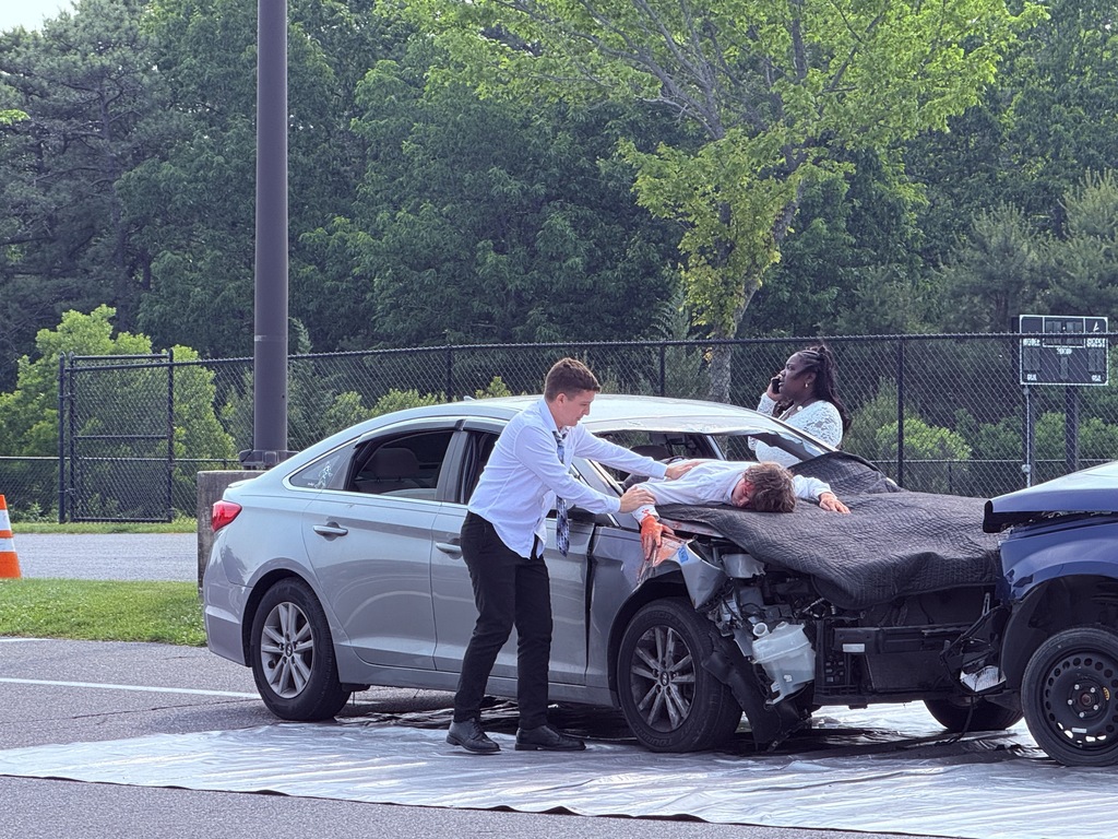 Scene of simulated car crash. There are three students in the picture, one is on the phone with 911, one is through the windshield laying on the hood of the car, and one is checking on the student on the hood of the car. 