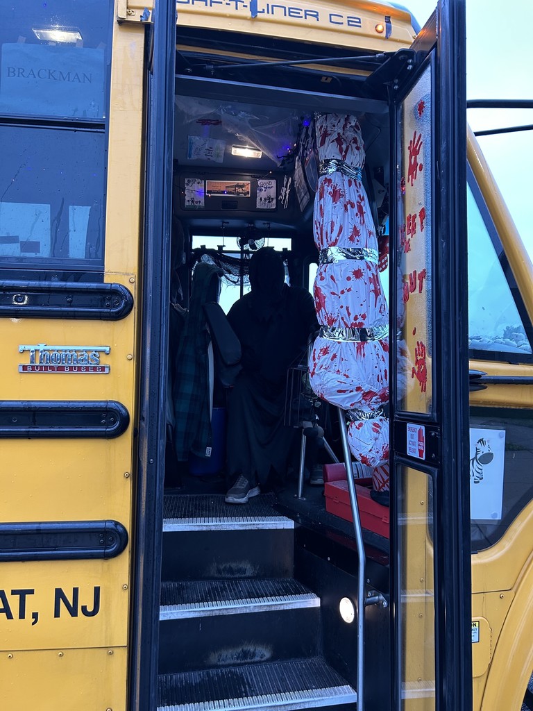 Image of the bus driver in costume with a halloween decoration in the entry way to the bus door. 