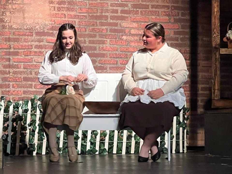 Image of two female actors sitting on a bench. 