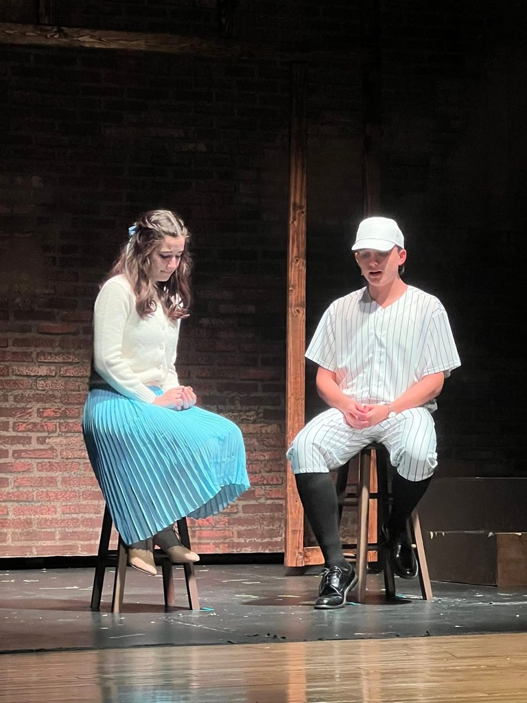 Image of a scene where the two actors are each sitting on a stool. 
