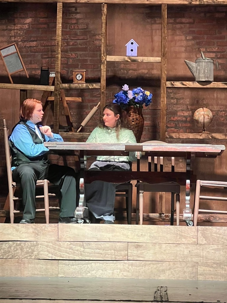 Image of a scene where the actors are sitting at a table. 