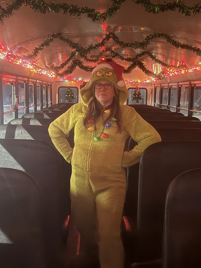 Image of the female bus driver standing in the aisle dressed up in a Grinch onesie with a santa hat and a light necklace.  