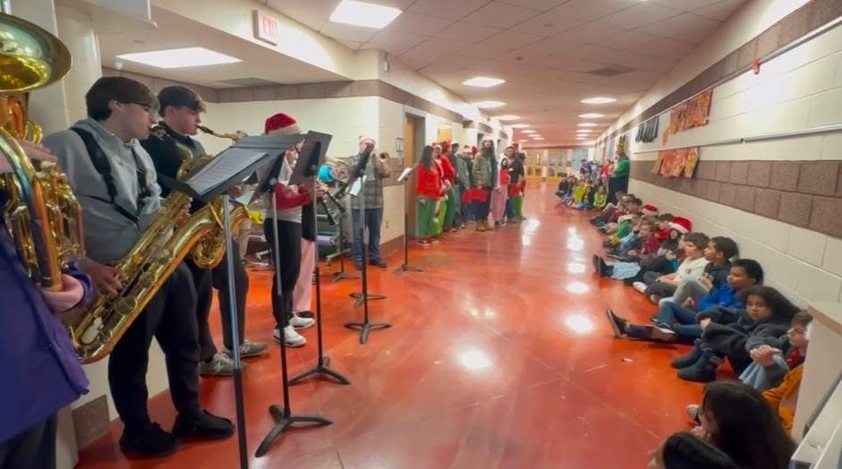 Image of members of the BHS band playing music in the hallway of the Donahue School while students sit against the wall to listen. 