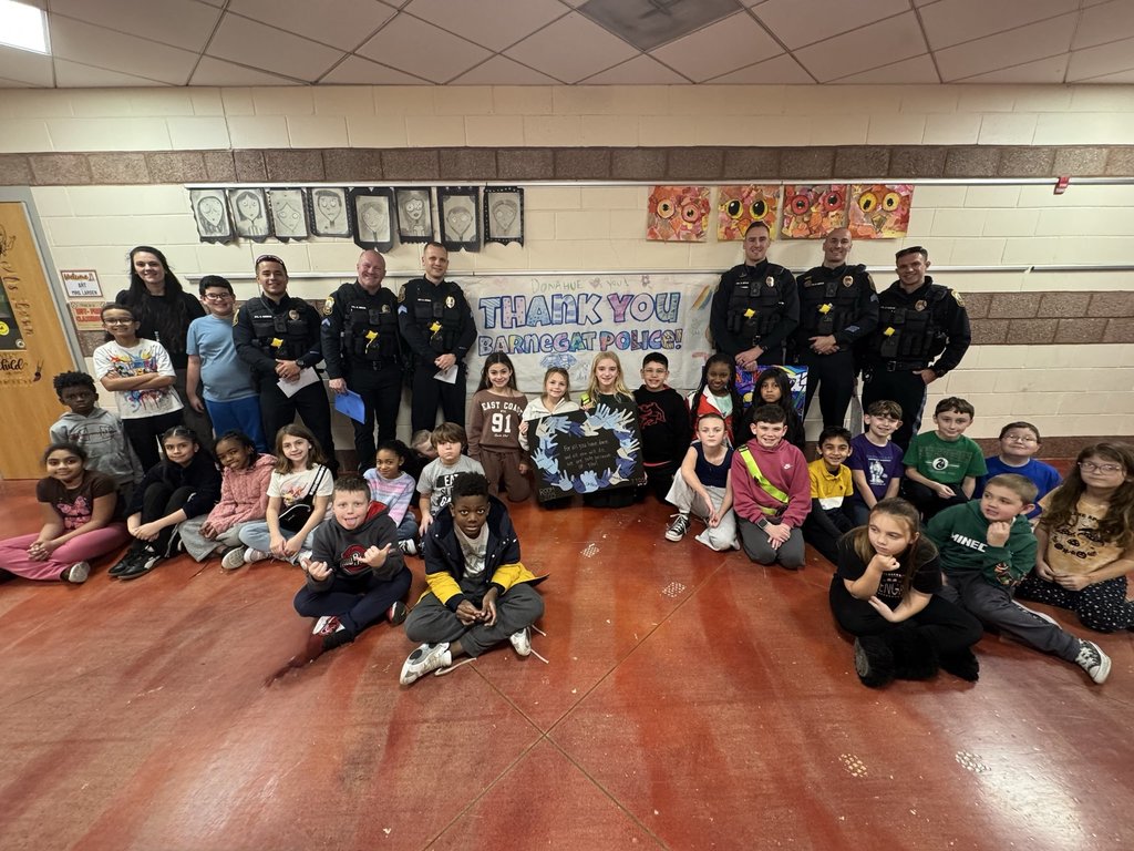 Image of a Donahue School class posing with six Barnegat police officers with a banner and a classroom handprint project thanking the police.  