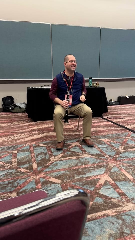 Image of Anthony Orrechio sitting in a chair holding a clarinet while presenting at NJMEA.  