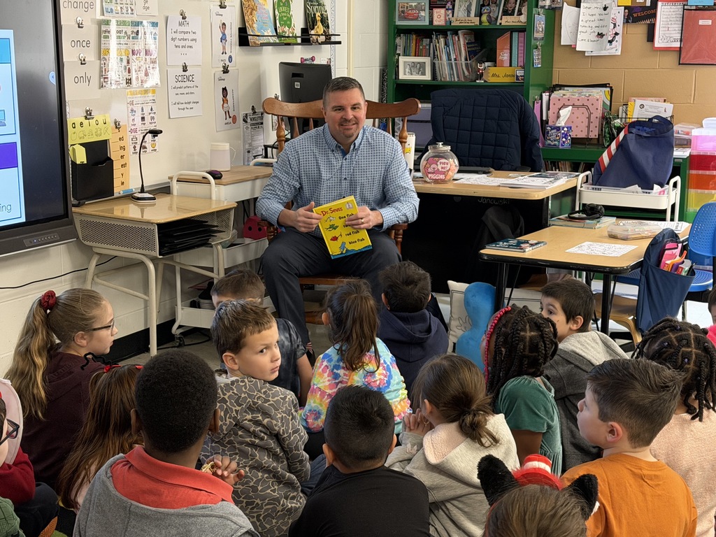 Image of a community member sitting in a chair holding a book getting ready to read to a class.