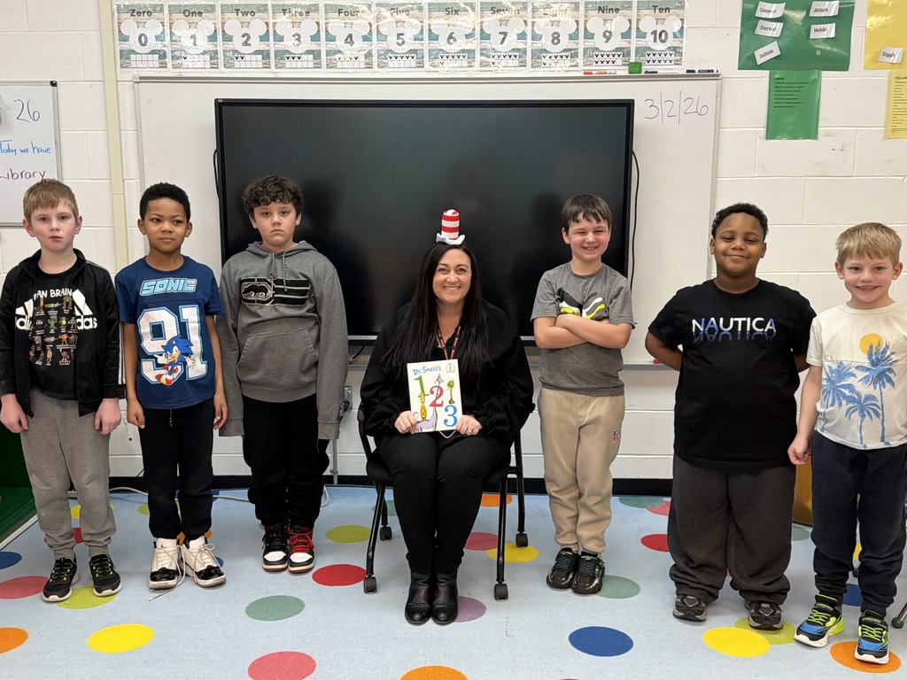 Image of a staff member sitting in a chair holding a book with three male students to her left and three male students to her right. 