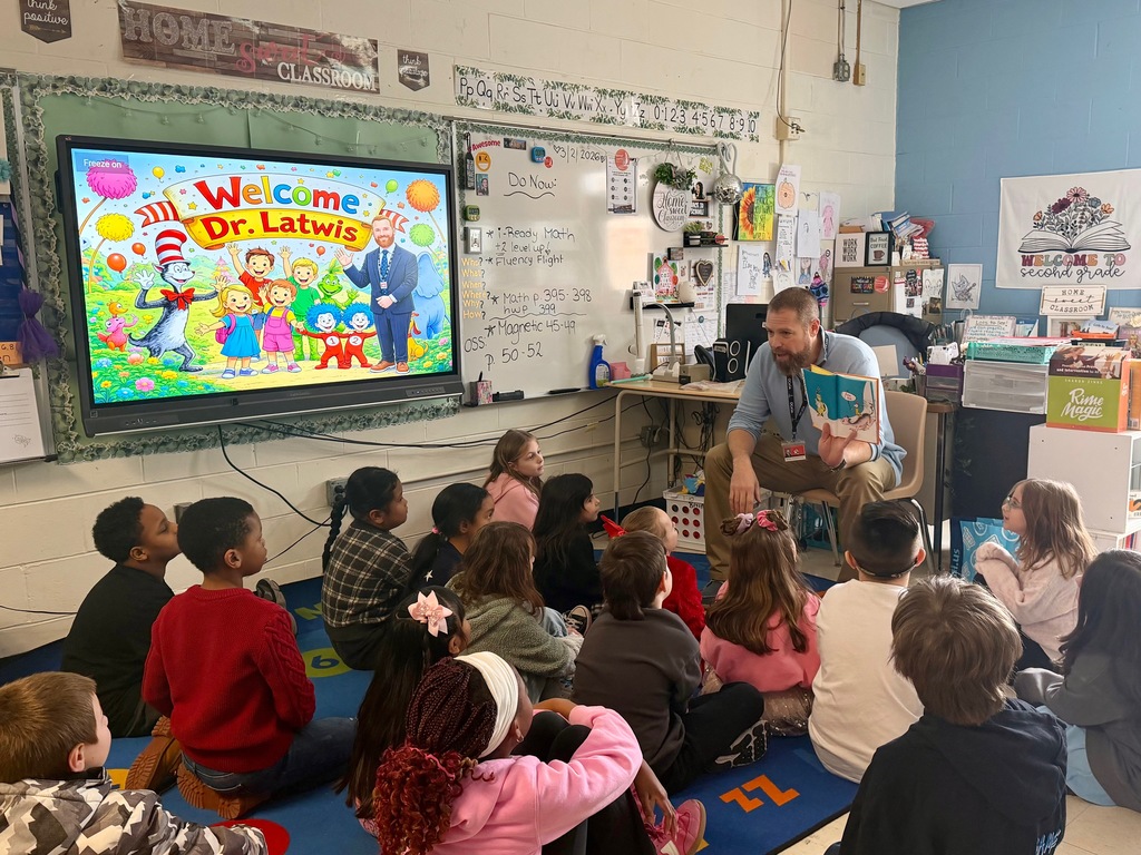 Image of the Superintendent sitting on a chair holding a book while he reads to a class. 