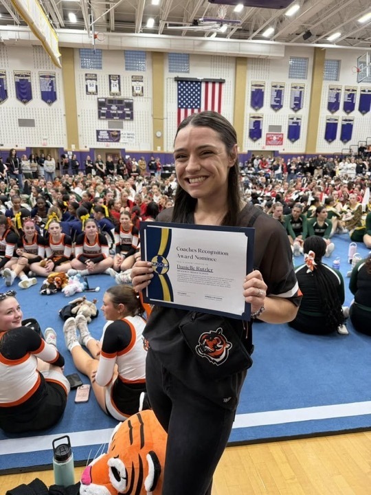 Image of Danielle holding the certificate she received with cheerleaders behind her on the mat. 