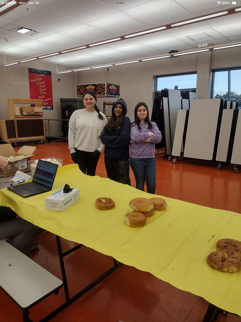 Three female students standing in front of the table full of bagels that they were helping distribute. 
