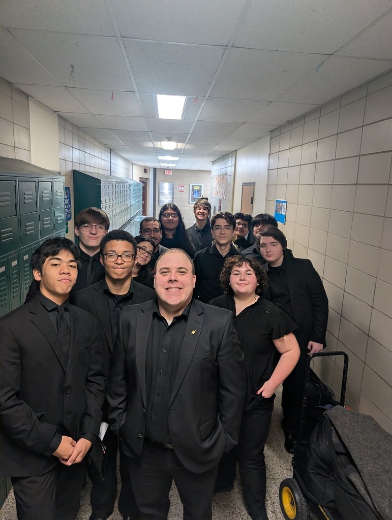Image of the Jazz Ensemble posing for a picture in the hallway of the school with the director. 