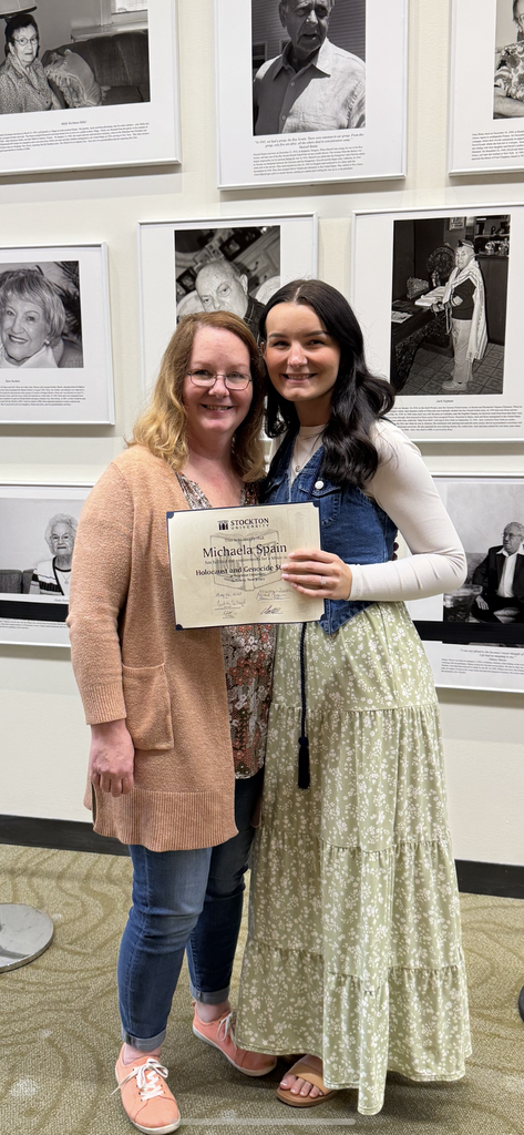 Image of Jill and Michaela posing together in front of a wall of Holocaust photos with Michaela holding a certificate. 