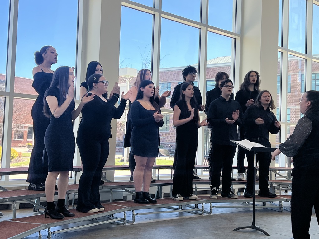 Image of the Select Choir performing on rafters in front of a wall of windows with their director facing them. 