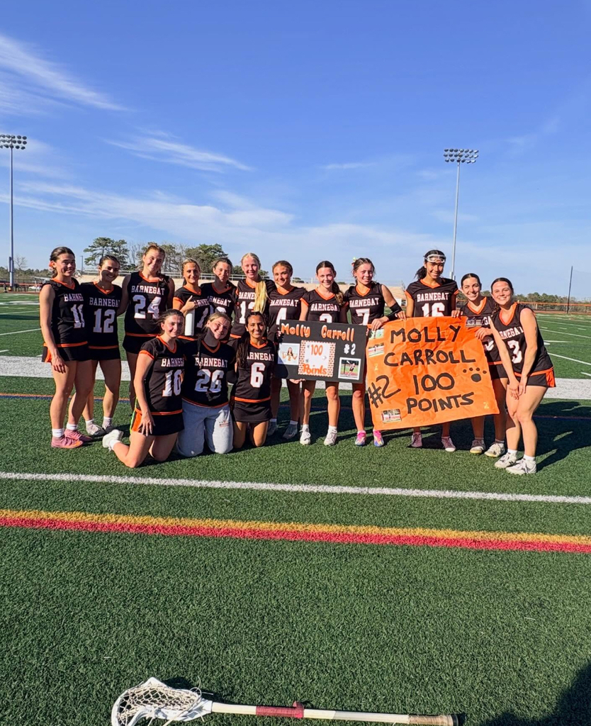 Image of Molly posing with the girls lacrosse team holding signs in support of her 100th point with her lacrosse stick in the foreground. 