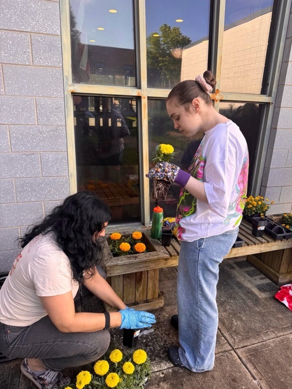 Image of two female students planting flowers in containers that are attached to a bench. 