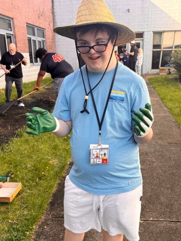 Image of a male MD student wearing garden gloves and a hat posing for a picture while a student and staff member are digging in the garden in the background. 
