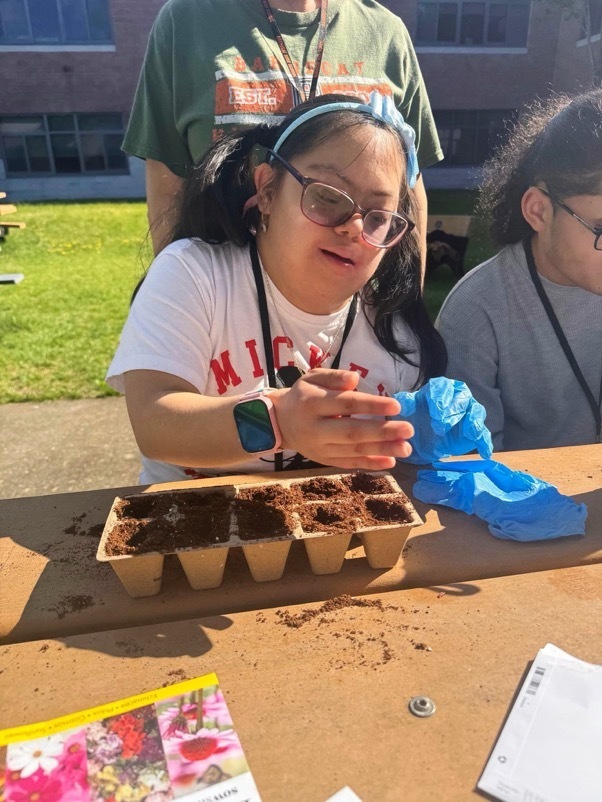 Image of a female MD student wearing gloves and planting seeds in soil in a planting container. 