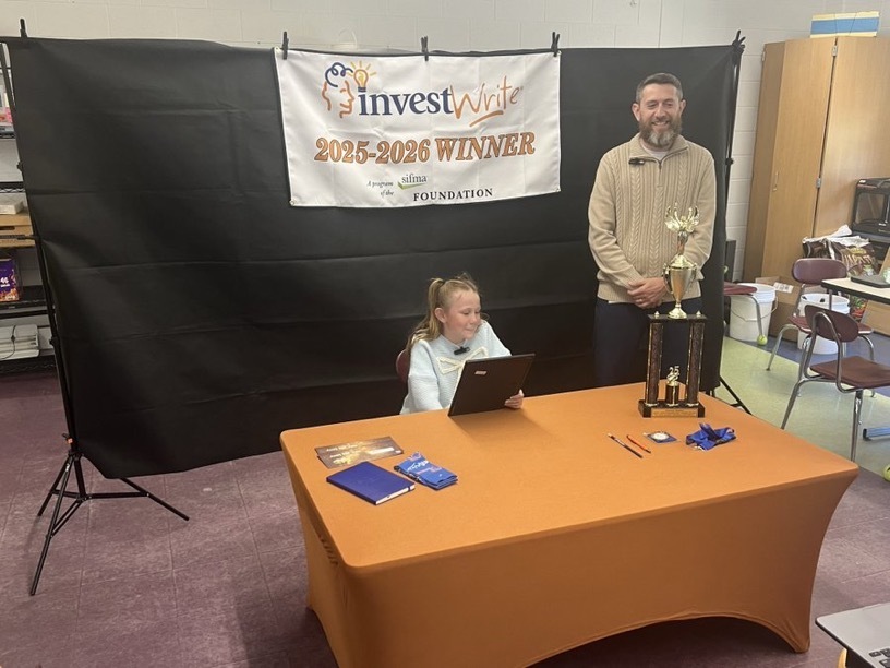 Image of Kennedy sitting at a table with her certificate and trophy and her teacher standing next to her. 