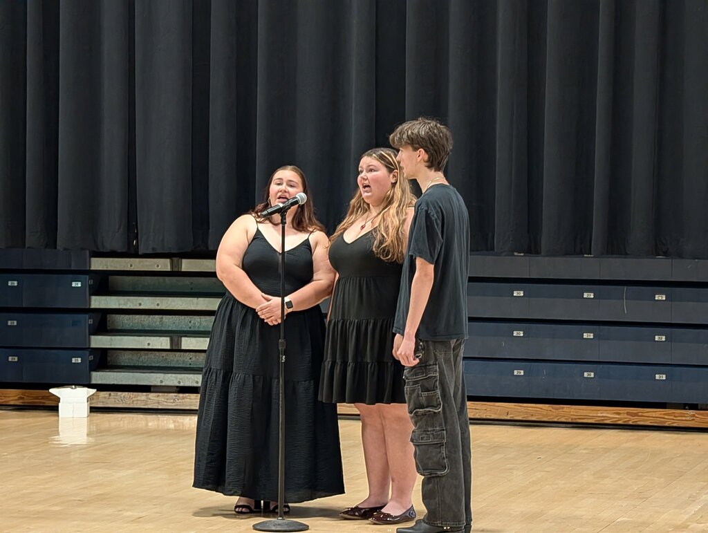 Image of the Select Choir members, two females and one male, singing into a microphone performing at the arena .   