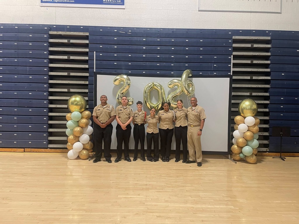 Image of the NJROTC members with Mr. Falcone posing in front of a picture wall that has ballon stacks to the left and right and 2026 balloons behind above their heads. 