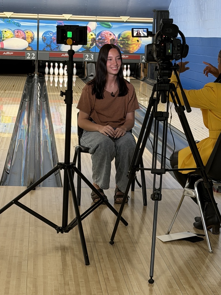 Image of Jamie sitting on a chair on a bowling lane with pins in the background and video equipment set up while she's being interviewed by a CBS rep.
