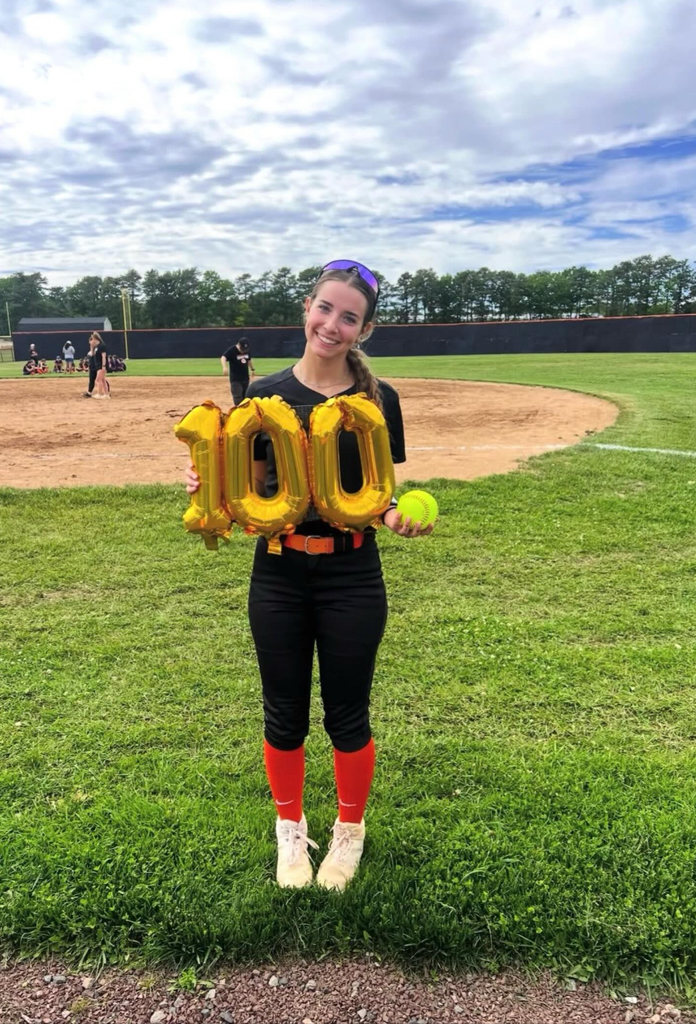 Image of Bri on the field holding a softball in one hand an balloons that read 100 in the other hand.