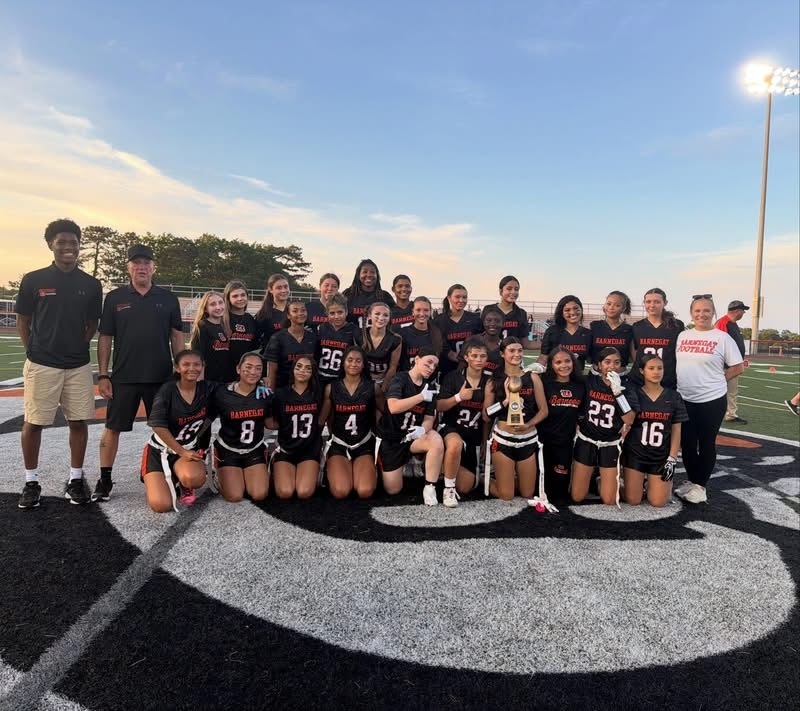 Image of the girls flag football team posing with the coaches on the field while one player holds the trophy after the championship win