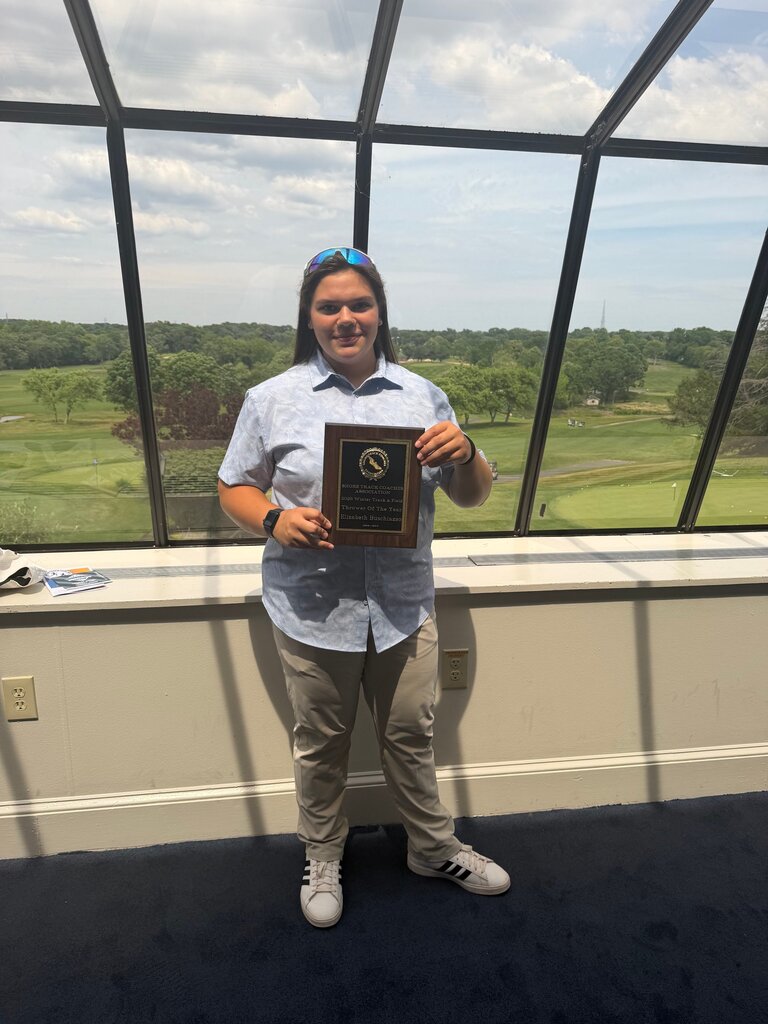 Image of Ellie holding a plaque in front of glass windows that look out on to a golf course. 