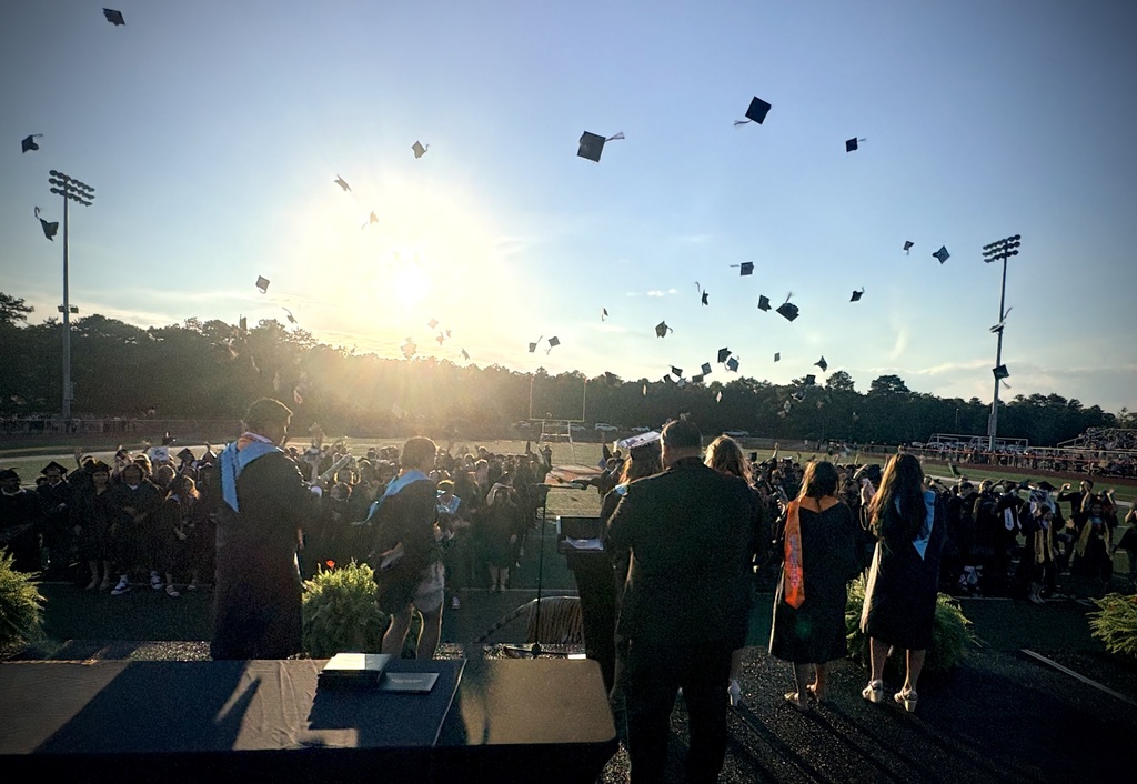 Image of the BHS Class of 2026 on the field throwing their graduation caps up in the air while the Principal and select students look out from the dais.