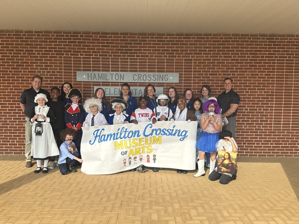 A group of children in costumes and several adults stand in front of Hamilton Crossing Elementary, holding a banner that reads “Hamilton Crossing Museum of Arts.”