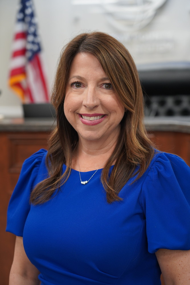 woman in blue dress standing in a board room next to a flag