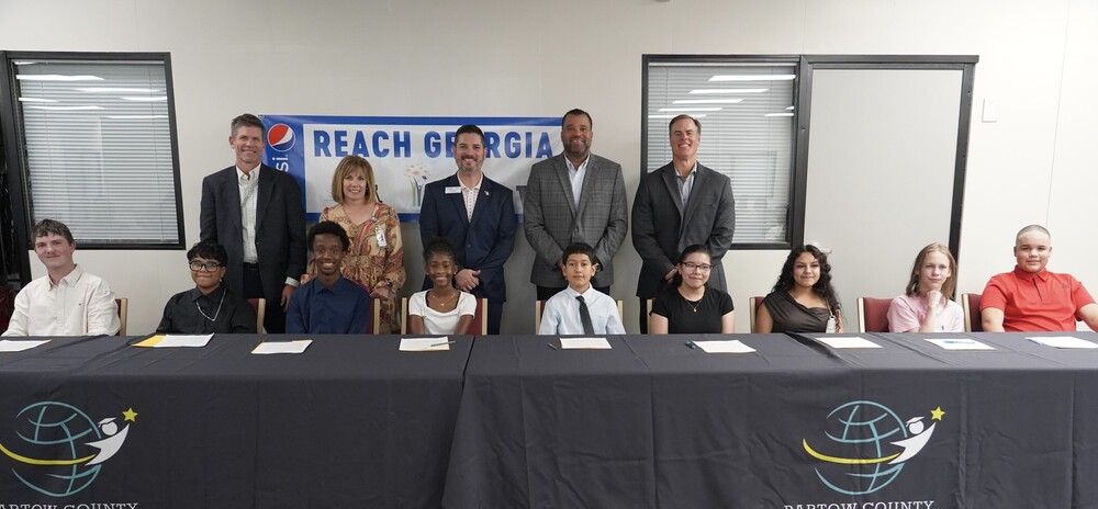Students sitting in a row with adults behind them signing a piece of paper