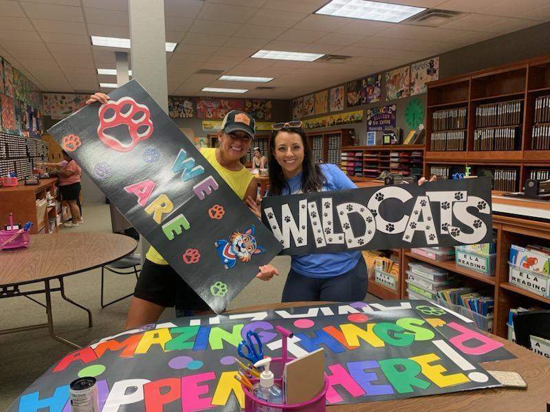 There are two teachers holding up signs. One says "We Are" and the other says "Wildcats".