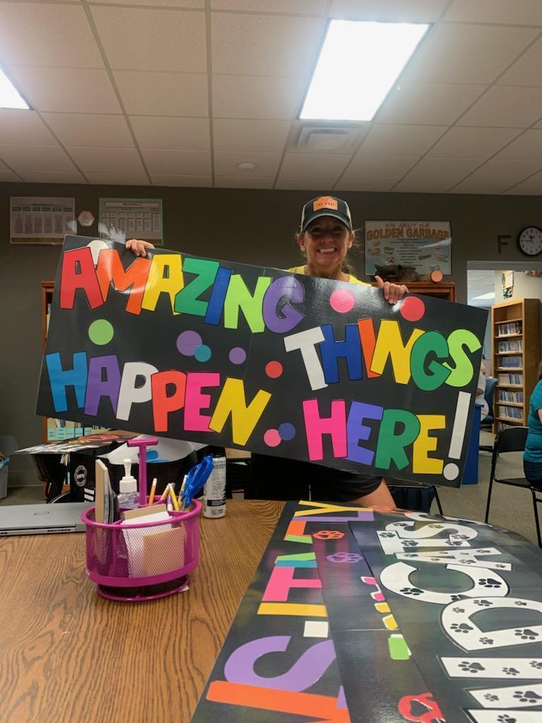 There is a teacher holding up a sign that says "Amazing Things Happen Here"!