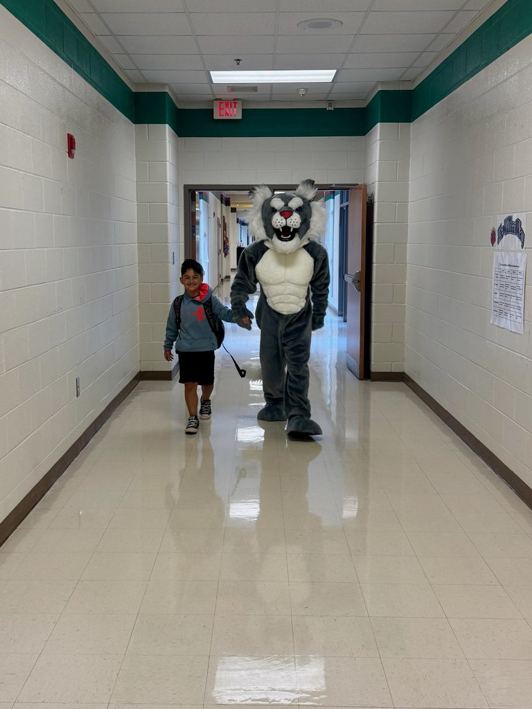 There is a young boy holding hands with Scratch, our Wildcat mascot, walking down the hall of the school.