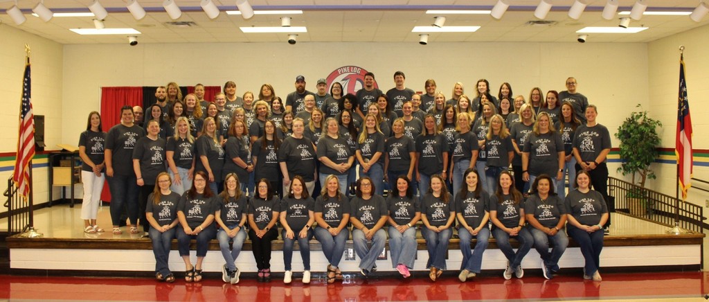 a group of 80 people all wearing a dark gray shirt and blue jeans sitting on a stage with an american flag on the left side and a georgia state flag on the right side. They are all smiling at the camera for a staff photo