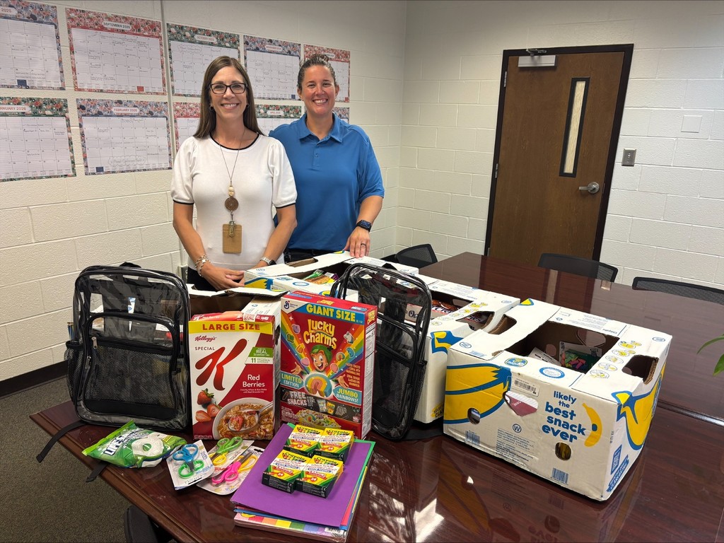 2 women standing in a conference room with calendars behind them and boxes of school supplies in front of them. There are cereal, crayons, backpacks and paper. The women are wearing a blue shirt and a white shirt