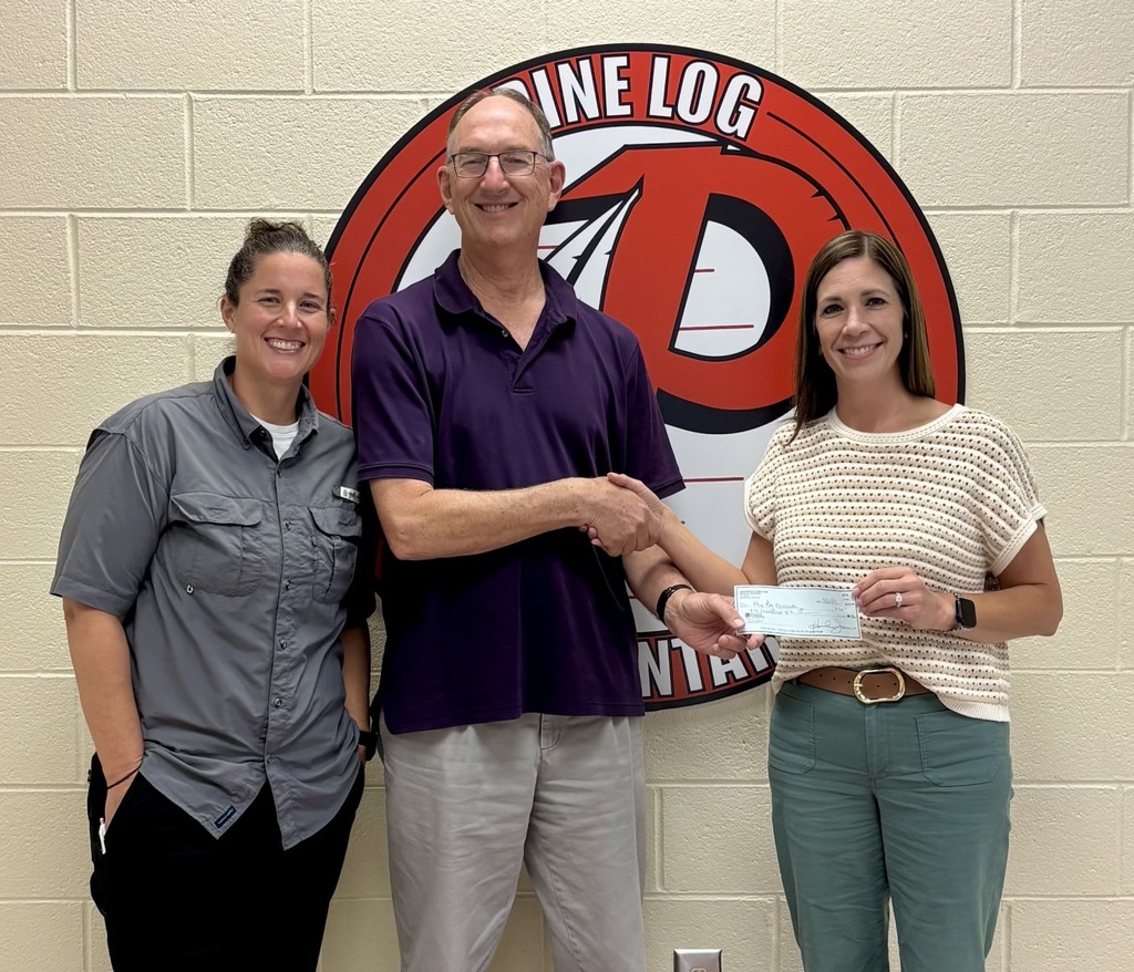 3 people standing in front of the Pine Log logo which is a circle with red and white writing and red P on it. The 2 women are dressed nice, one in green pants and a sweater shirt,  and the other in a blue button up dress shirt. The man is tall and is standing in the middle with a purple polo shirt on . 