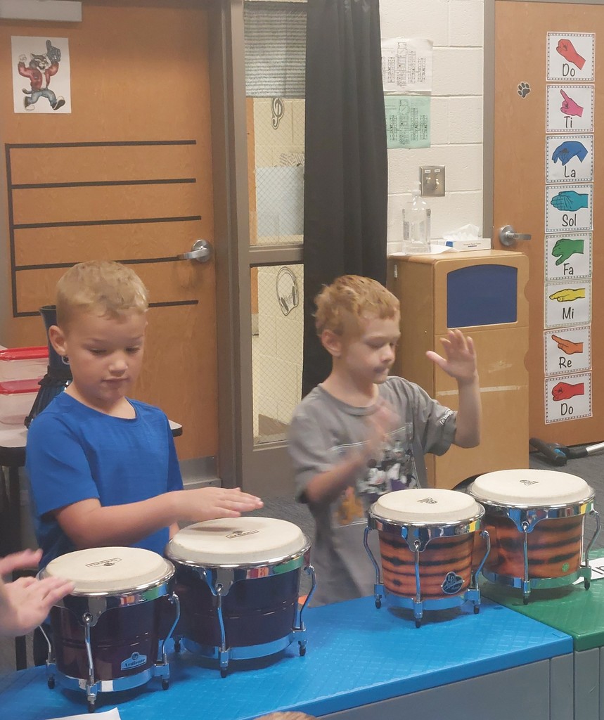 Two boys are playing the drums.  One is wearing a blue shirt and the other is wearing a grey shirt.