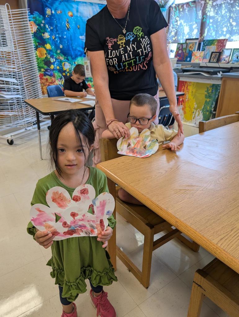 There is a girl in a green dress holding up a paw print that has been finger painted.  Behind her is a boy at the table with his paw print.