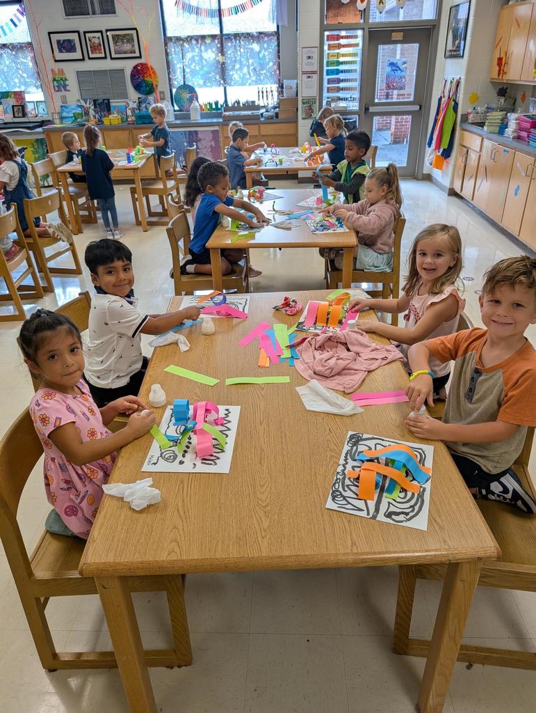 There is a table with two girls and two boys at it.  They are gluing sheets of construction paper to their paper to make a picture.  There are four other tables with boys and girls at them in the background.