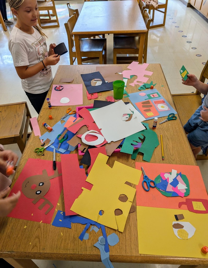 There is a table full of construction that has been ripped and glued on other sheets to make pictures.  There is one picture of a reindeer, one of ajar.  There is a girl standing there working on her picture.